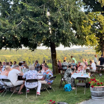 L'image représente un groupe de personnes réunies en plein air, probablement à l'occasion d'un pique-nique ou d'un événement en plein air, avec des tables et des chaises visibles à l'arrière-plan, ce qui donne un sentiment de communauté et de rassemblement social.