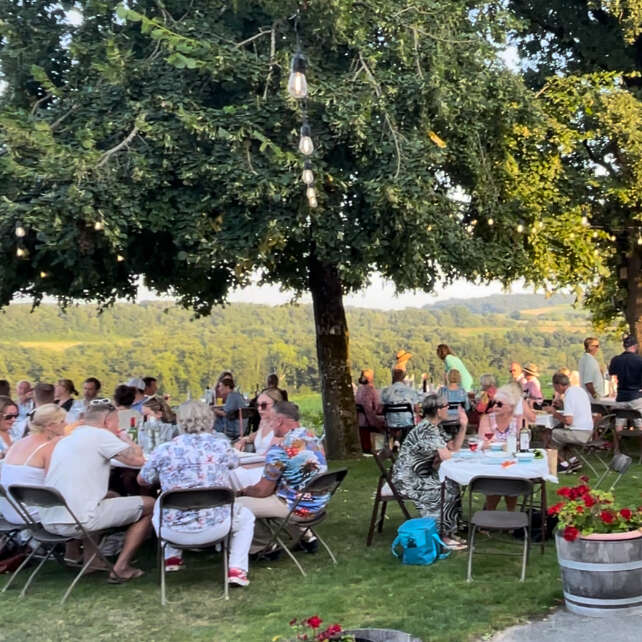 L'image représente un groupe de personnes réunies en plein air, probablement à l'occasion d'un pique-nique ou d'un événement en plein air, avec des tables et des chaises visibles à l'arrière-plan, ce qui donne un sentiment de communauté et de rassemblement social.