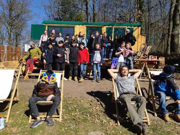 A large group of children and adults gather outdoors in front of a wooden cabin. Some relax on deck chairs, while others stand together, enjoying sunny weather.