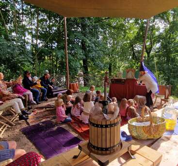 Children and adults sit outdoors on benches and mats watching a performer wearing a wizard hat. The area is shaded, surrounded by green trees, and decorated festively.