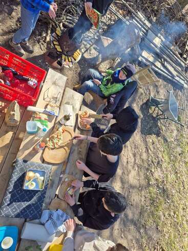 A group of people prepares food outdoors around a wooden table. Some are chopping vegetables, while others cook corn and pizza on a small fire grill.