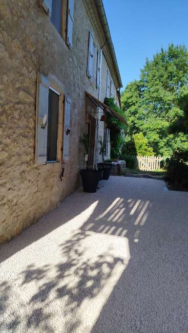A rustic stone house with pale blue shutters casts strong shadows onto a gravel path. Large potted plants and green trees surround the peaceful, sunlit setting.