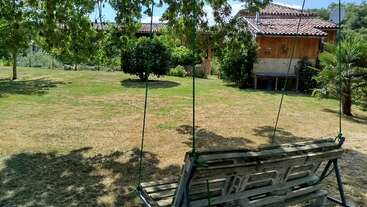 This image shows a peaceful garden with a wooden swing in the foreground, lush green trees, grassy lawn, and a rustic house in the background.