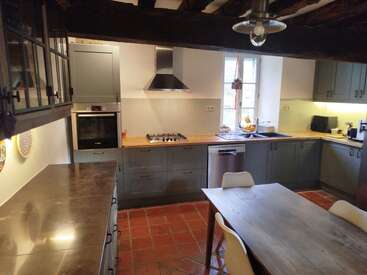 This kitchen features rustic wooden beams, grey cabinets, stainless steel appliances, a wooden dining table, white chairs, tiled floor, and ample natural light from a window.