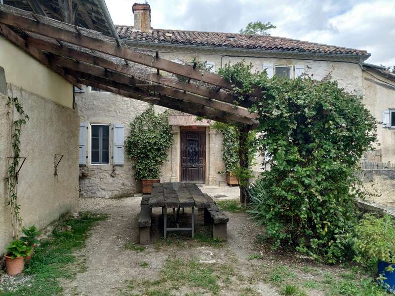 A rustic stone house with wooden table and benches outdoors. Wooden pergola covered in vines, plants around, blue shutters, and a charming countryside atmosphere.
