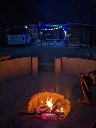 A cozy night scene with a campfire burning in a circular pit, a person sitting nearby, and colorful string lights illuminating a rustic outdoor shelter.