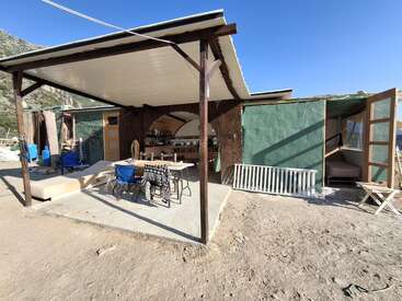 A rustic outdoor kitchen and seating area with simple furniture, surrounded by unfinished walls, under a covered patio, set in a rural, mountainous environment.