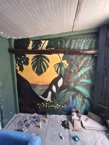 A vibrant mural features lush tropical plants, fern leaves, and flowers framing a sunset landscape. Painting materials and supplies are scattered on the floor, indicating ongoing work.