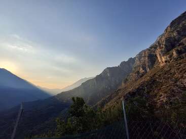 A breathtaking mountain landscape at sunset, with rugged cliffs, lush greenery, and a serene sky fading from golden light to a deep blue, separated by a fence.