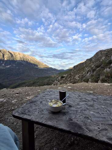 A rustic wooden table holds a bowl of cereal and a glass of coffee, overlooking stunning mountains, dramatic sky, and serene valley under soft morning light.