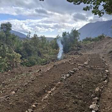 A freshly prepared garden bed divided by stones sits on a hillside. Smoke rises in the background, surrounded by trees and distant mountains under a cloudy sky.
