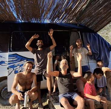 A group of people, including children and adults, happily pose and wave in front of a van under a bamboo canopy, enjoying a relaxed, sunny outdoor moment.