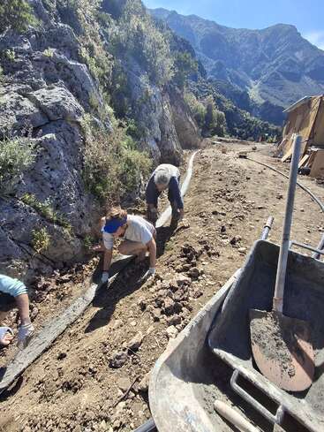 Dos personas trabajan en un camino de tierra cerca de un terreno rocoso, utilizando hormigón y herramientas, rodeadas de montañas y naturaleza bajo un cielo azul y despejado, dando forma a un bordillo.