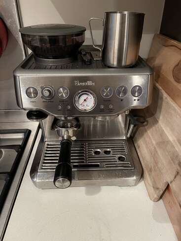 La imagen muestra una cafetera espresso Breville de acero inoxidable sobre una encimera de cocina, con un molinillo de café y un espumador de leche encima.