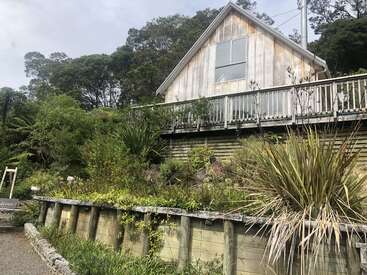 La imagen muestra una casa de madera desgastada con un balcón, rodeada de exuberante vegetación y un muro de piedra, bajo un cielo nublado.