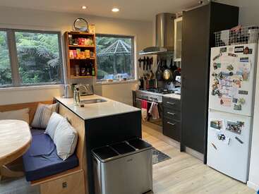 La imagen muestra una cocina moderna con zona de comedor, que incluye un frigorífico blanco, armarios oscuros y una ventana con vistas a un frondoso jardín.