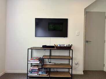 La imagen muestra una habitación minimalista con un televisor montado en la pared sobre una estantería, con libros y una cesta de objetos en los estantes.