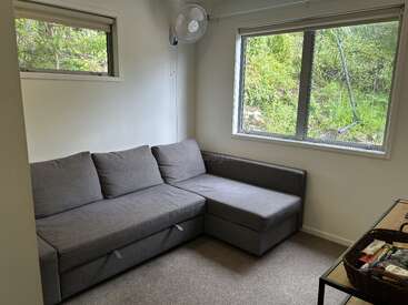 La imagen muestra una acogedora habitación con un sofá gris, dos ventanas y un ventilador, con una serena vista de la exuberante vegetación exterior.