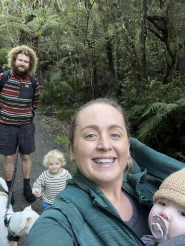 Una familia sonriente pasea por un frondoso sendero forestal. Una persona sostiene a un bebé con chupete, mientras otra pasea a un perro junto a un niño pequeño.
