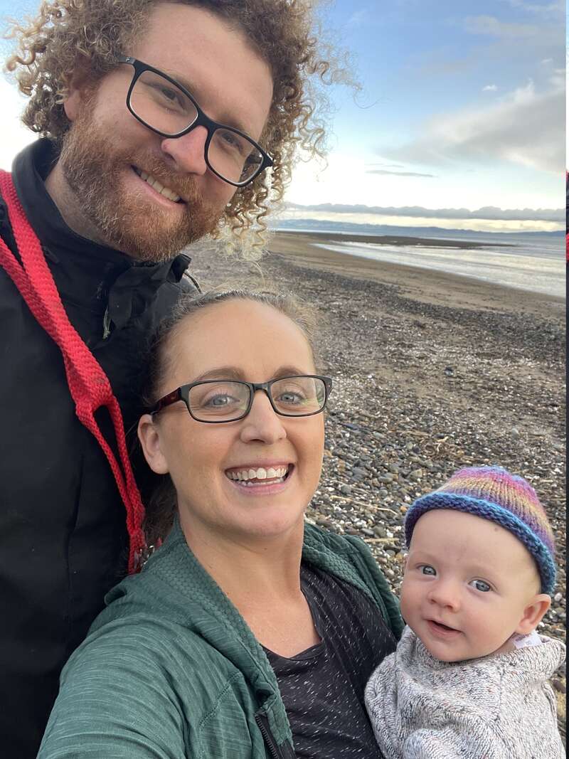 La imagen muestra a una familia de tres miembros posando para un selfie en una playa, con el hombre y la mujer de pie detrás del bebé, que lleva un colorido gorro de punto.