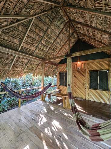 This rustic wooden cabin features a thatched roof, two colorful hammocks, a wooden bench, and large windows, surrounded by lush green tropical vegetation, inviting relaxation.