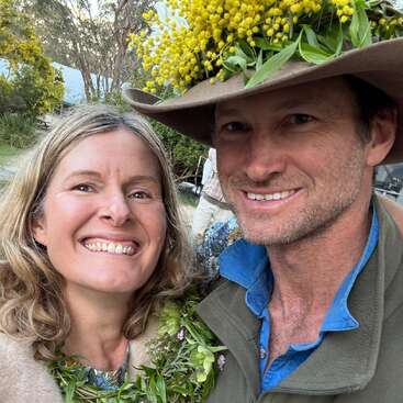 Eine Frau und ein Mann lächeln und posieren draußen. Der Mann hat einen Hut mit gelben Blumen und Blättern drauf, und beide haben grünes Laub um den Hals.