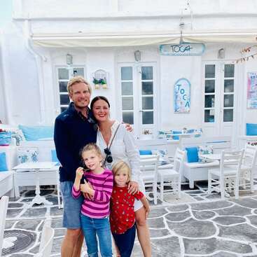 The image depicts a family of four posing for a photo in front of a white building with blue accents, likely a restaurant or cafe.