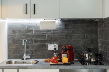 This kitchen features a sleek sink, modern tap, dark subway tile backsplash, red coffee machine, vegetables on a cutting board, pot on stove, and under-cabinet lighting.