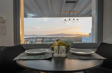 A modern dining table set for two overlooks a beautiful sunset view through a large window, featuring fruit, salt, pepper, and stylish decor.