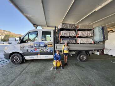 Two children in sailing gear pose beside a truck loaded with covered sailing equipment. The truck displays sailing-themed graphics and is parked under a large shelter.