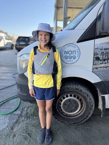 A smiling child in sailing gear and a bucket hat stands next to a van with Greek writing, ready for an outdoor adventure on a sunny day.