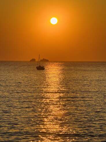 A golden sunset bathes the calm sea in warm light, with a tranquil sailboat gently floating and distant rocks silhouetted against the glowing horizon. Peaceful serenity.