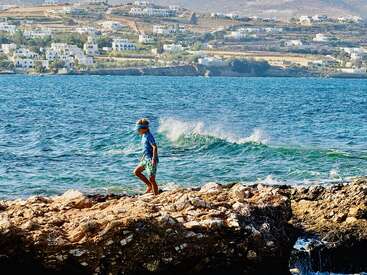 A boy walks barefoot on rocky shore by the sea, with turquoise waves behind him and white houses dotting the hillside under a bright, sunny sky.