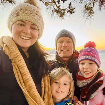 A smiling family of four, dressed warmly in hats and scarves, enjoys an outdoor winter day together with a scenic sunset and tree branches in the background.
