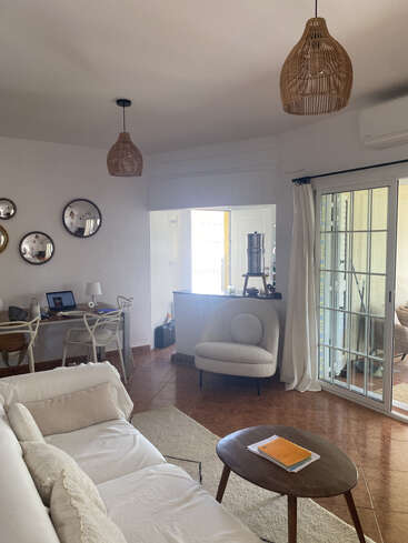 Un salon confortable et moderne avec un canapé blanc, un fauteuil rond, une table basse en bois, des suspensions lumineuses, des miroirs, un bureau avec un ordinateur portable et des portes coulissantes en verre.