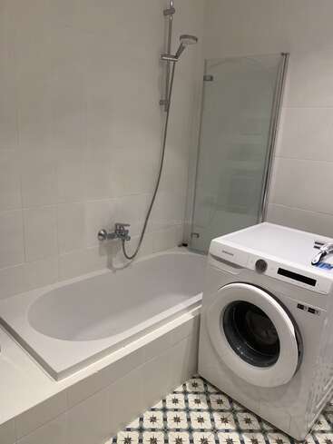 Cette image montre une salle de bain moderne équipée d'une baignoire, d'une douche, d'une cloison en verre et d'un lave-linge Samsung. Le sol est recouvert de carreaux à motifs bleus et blancs.
