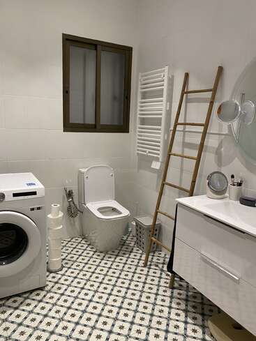 Cette salle de bain est équipée d'un carrelage à motifs, d'un lave-linge, de toilettes, d'un porte-serviettes, d'une échelle en bambou, d'un meuble-lavabo avec miroir circulaire et d'une fenêtre dépolie. Élégant et moderne.