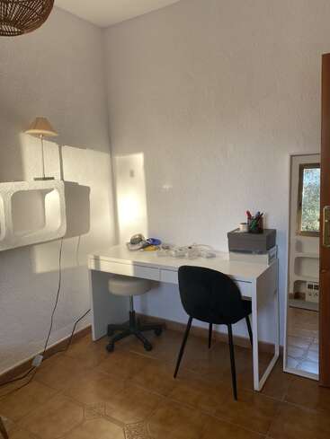 Cette image montre une petite pièce ensoleillée avec un bureau blanc, deux chaises, une lampe, un sol carrelé, des murs texturés et des fournitures de bureau soigneusement rangées.