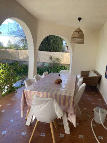 Une terrasse couverte avec des ouvertures en arc est équipée d'une grande table recouverte d'une nappe rayée, de chaises blanches, d'un fauteuil en osier, de plantes, d'un sol carrelé et d'un luminaire suspendu.