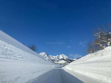 The image depicts a snow-covered road with tire tracks, flanked by snowbanks and trees, set against a backdrop of mountains under a clear blue sky.