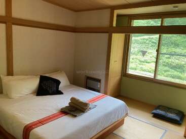 Das Bild zeigt ein ruhiges Schlafzimmer mit einem Doppelbett, weißer Bettwäsche und einem Fenster, das den Blick auf einen üppig grünen Berghang freigibt.