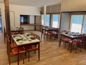 Das Bild zeigt einen geräumigen Raum mit mehreren Tischen und Stühlen, Holzböden und -wänden und großen Fenstern, in dem eine Mahlzeit mit einer Auswahl an Tellern und Schüsseln serviert wird.