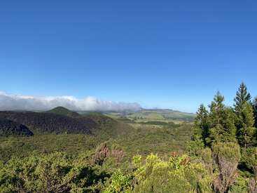 La imagen representa un paisaje sereno de exuberante vegetación, colinas ondulantes y un cielo azul despejado, con una cordillera lejana parcialmente oculta por las nubes.