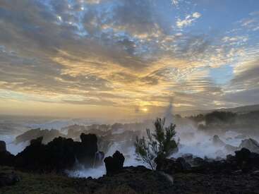La imagen representa una impresionante puesta de sol sobre una costa escarpada, con las olas rompiendo contra las rocas y un cielo espectacular lleno de nubes.