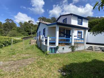 Una encantadora casa blanca con detalles azules se encuentra en un exuberante jardín verde. Rodeada de árboles, tiene un porche, escalones y un cielo azul despejado.