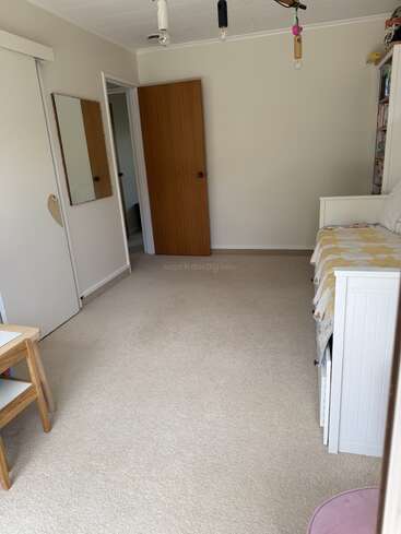 A tidy, beige-carpeted room with white walls, open wooden door, mirror, small table, bench seat with quilt, bookshelves, and exposed ceiling lights. Simple decor.