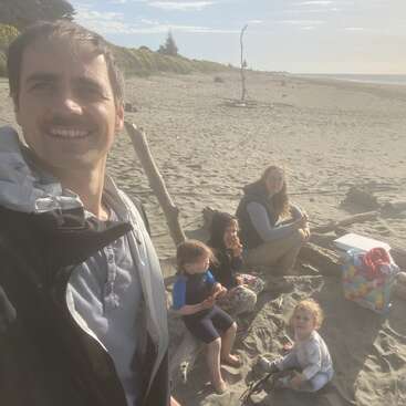 A family enjoys a sunny day at the beach. The father takes a selfie while the mother and three children sit together on the sand, smiling.