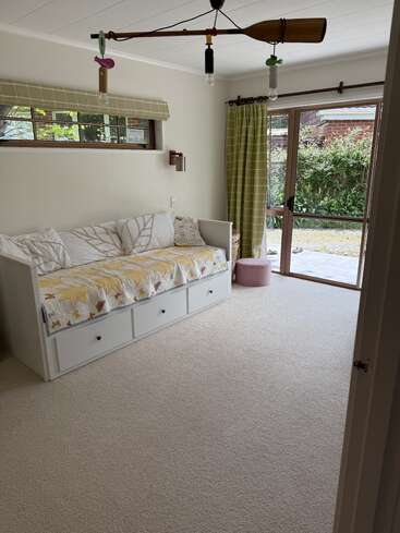 A bright, cozy room with a daybed featuring leaf-patterned bedding, storage drawers beneath, green curtains, large window doors, and a paddle-shaped ceiling light fixture.