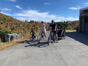 A family of three, wearing helmets, rides bikes on a sunny day. The child leads, parents follow, with a trailer attached. Hills and blue sky background.