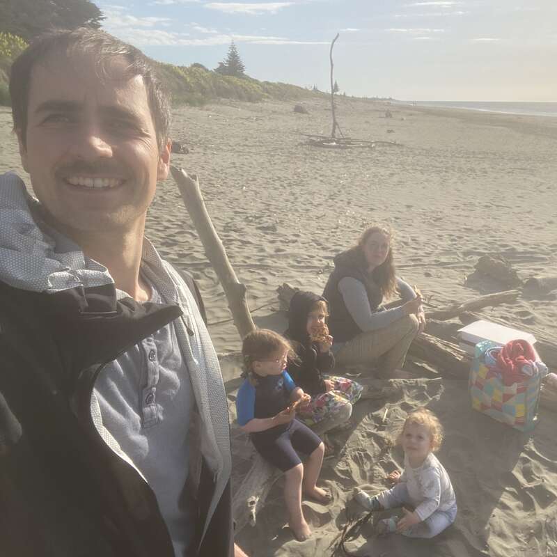 A family enjoys a sunny day at the beach. The father takes a selfie while the mother and three children sit together on the sand, smiling.